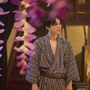 Person in a patterned kimono standing indoors, with pink flower decorations hanging in the foreground and a traditional lamp in the background.