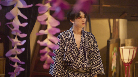 Person in a patterned kimono standing indoors, with pink flower decorations hanging in the foreground and a traditional lamp in the background.