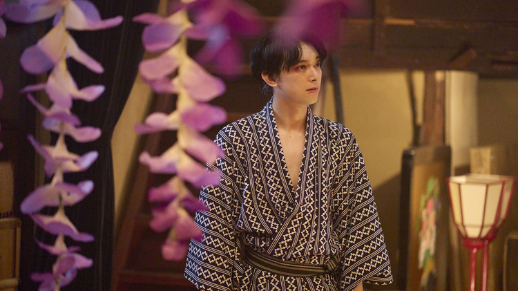 Person in a patterned kimono standing indoors, with pink flower decorations hanging in the foreground and a traditional lamp in the background.