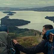 Two hikers with backpacks overlooking a large lake and islands from a grassy hilltop.
