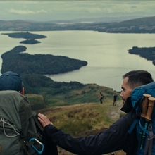 Two hikers with backpacks overlooking a large lake and islands from a grassy hilltop.