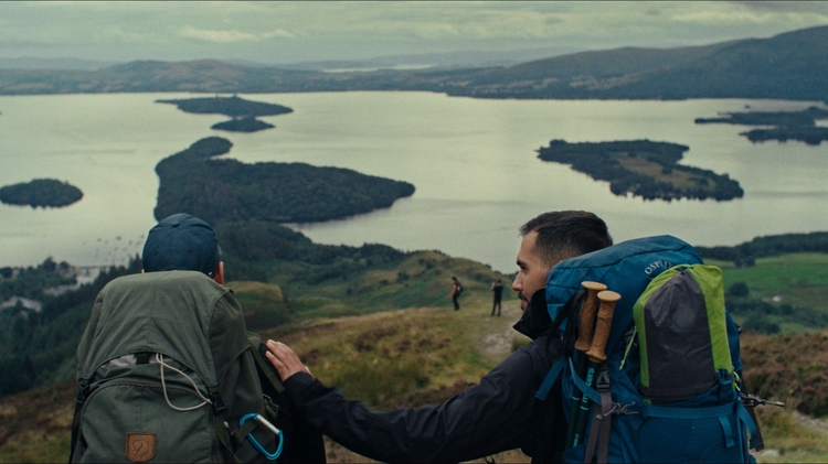 Two hikers with backpacks overlooking a large lake and islands from a grassy hilltop.