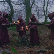 Five robed figures in a forest circle a small fire, each holding a flaming torch pointing toward the centre pile of sticks.