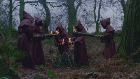 Five robed figures in a forest circle a small fire, each holding a flaming torch pointing toward the centre pile of sticks.