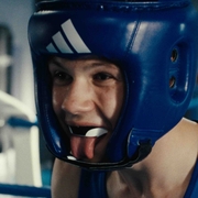 Boxer wearing a blue headguard and a blue tank top, standing inside a boxing ring with blurred gym equipment in the background.
