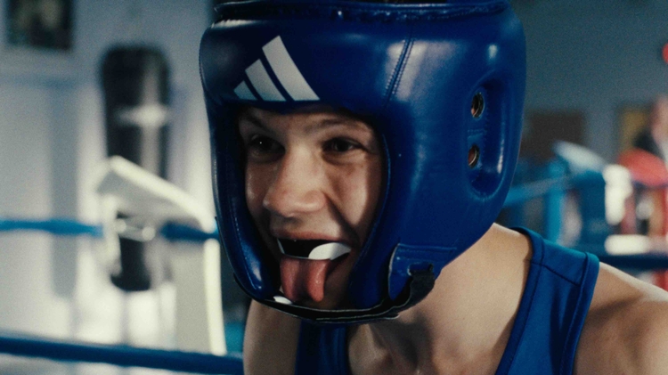 Boxer wearing a blue headguard and a blue tank top, standing inside a boxing ring with blurred gym equipment in the background.