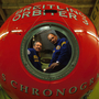 Two people in blue uniforms inside a large red Breitling Orbiter 3 capsule in a wooden indoor setting.