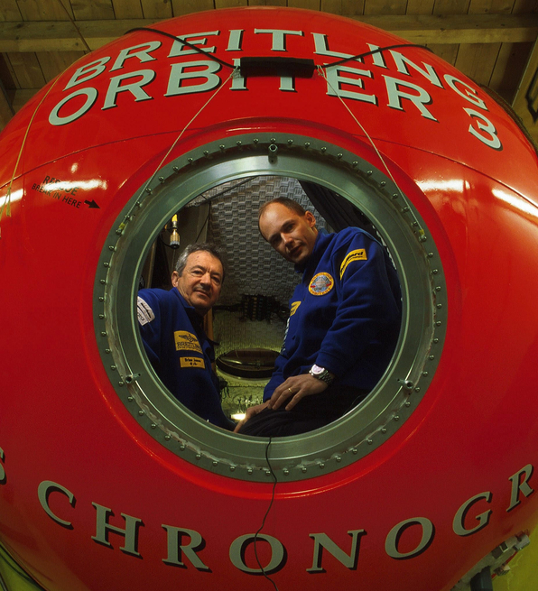 Two people in blue uniforms inside a large red Breitling Orbiter 3 capsule in a wooden indoor setting.