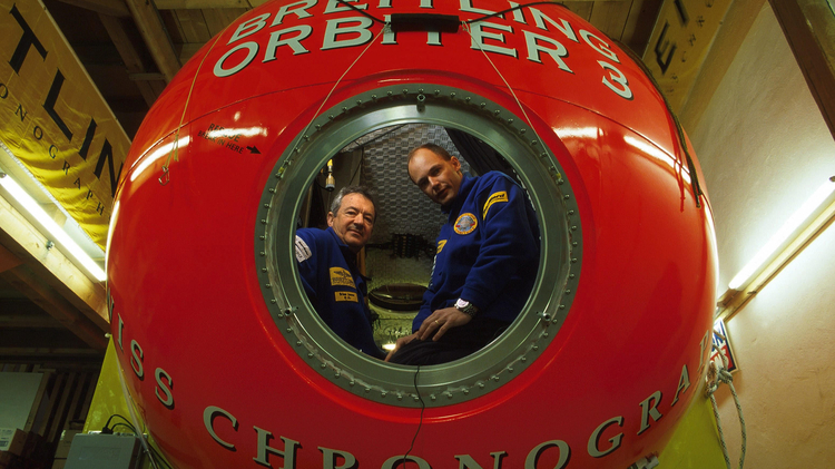 Two people in blue uniforms inside a large red Breitling Orbiter 3 capsule in a wooden indoor setting.