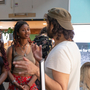 Three Black and Brown people engaged in conversation at an indoor event. Two women listen attentively to a man gesturing as he speaks. The setting is casual and lively.
