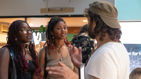 Three Black and Brown people engaged in conversation at an indoor event. Two women listen attentively to a man gesturing as he speaks. The setting is casual and lively.
