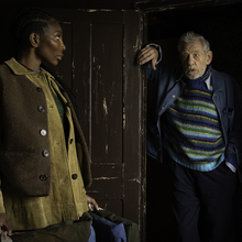 Two people standing indoors near a worn wooden door; one leans on the doorframe, the other holds a bag, both dressed in layered clothing.