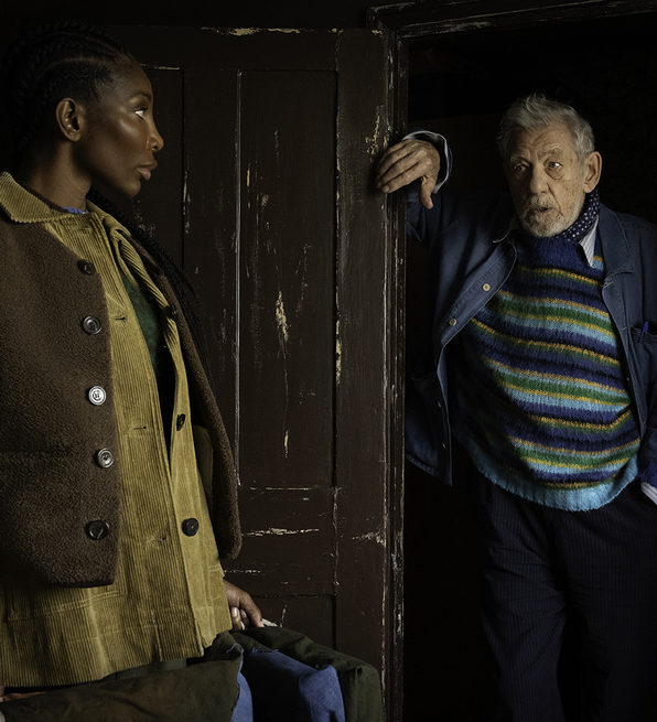 Two people standing indoors near a worn wooden door; one leans on the doorframe, the other holds a bag, both dressed in layered clothing.
