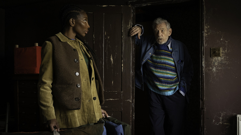Two people standing indoors near a worn wooden door; one leans on the doorframe, the other holds a bag, both dressed in layered clothing.