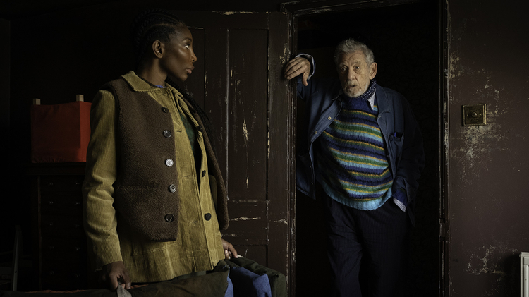 Two people standing indoors near a worn wooden door; one leans on the doorframe, the other holds a bag, both dressed in layered clothing.