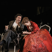 Two people in formal attire sit on chairs in a dark setting, holding glasses, one wearing a red embellished gown.