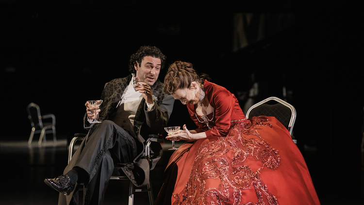 Two people in formal attire sit on chairs in a dark setting, holding glasses, one wearing a red embellished gown.