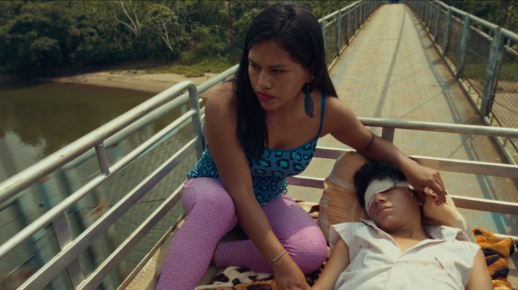 Two people sitting on a truck bed, one sitting upright and comforting the other lying down on a blanket.