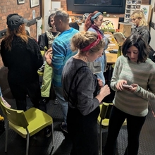 A group of people in a cozy, dimly-lit room with brick walls, illuminated by string lights. They chat and engage in activities around a table. Relaxed atmosphere.