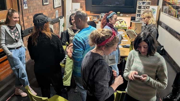 A group of people in a cozy, dimly-lit room with brick walls, illuminated by string lights. They chat and engage in activities around a table. Relaxed atmosphere.