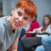 A white person with short red hair and gold hoops leans forward, holding a pen, appearing engaged. They are in a casual setting with others blurred in the background.