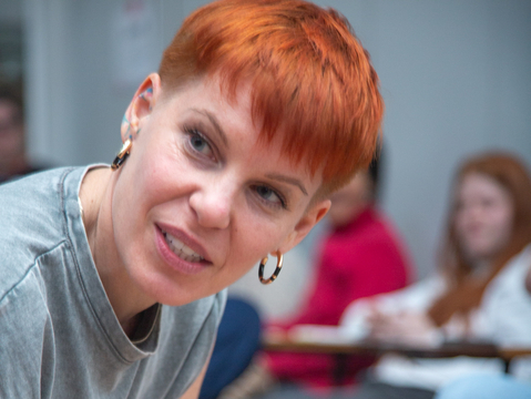 A white person with short red hair and gold hoops leans forward, holding a pen, appearing engaged. They are in a casual setting with others blurred in the background.