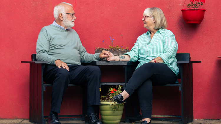 An elderly couple sit together holding hands, smiling at one another