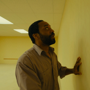 A still from Backrooms of a man in a brown shirt touching a plain yellow wall in an empty, fluorescent-lit room.