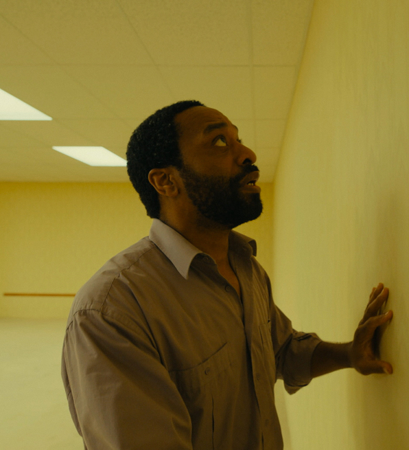 A still from Backrooms of a man in a brown shirt touching a plain yellow wall in an empty, fluorescent-lit room.