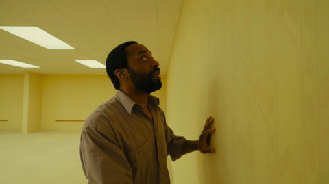 A still from Backrooms of a man in a brown shirt touching a plain yellow wall in an empty, fluorescent-lit room.