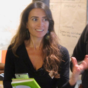 Two women are engaged in discussion indoors, one gesturing with enthusiasm. A beige flip chart with notes is visible in the background.