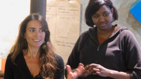 Two women are engaged in discussion indoors, one gesturing with enthusiasm. A beige flip chart with notes is visible in the background.