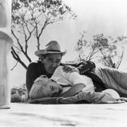 Black and white photo of a cowboy in a hat lying on the ground, holding a resting blonde woman outdoors near trees.