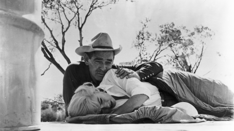 Black and white photo of a cowboy in a hat lying on the ground, holding a resting blonde woman outdoors near trees.