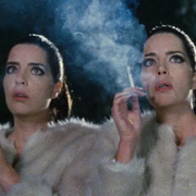 A still from Diamantino of two women in white fur coats smoking cigarettes outdoors at night with smoke visible around them.