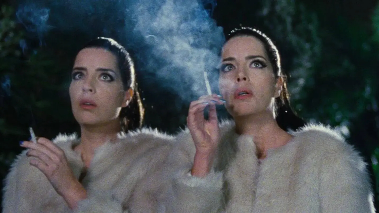A still from Diamantino of two women in white fur coats smoking cigarettes outdoors at night with smoke visible around them.