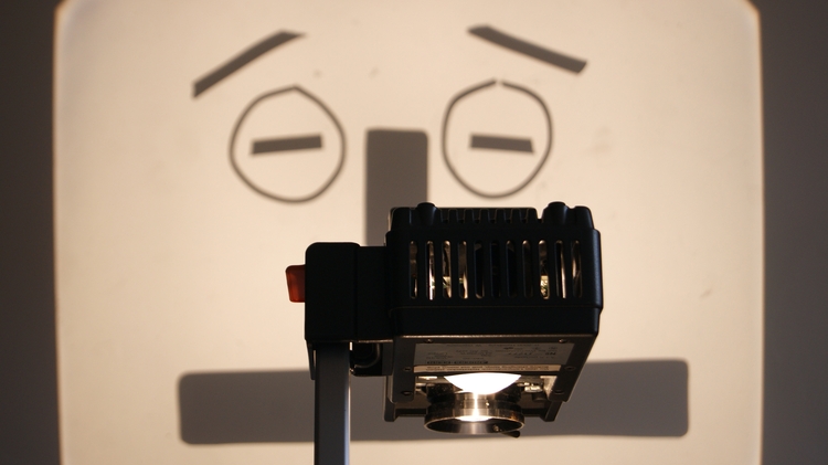 An overhead projector casts a shadow resembling a surprised face with raised eyebrows and round eyes. The mood is playful and creative.