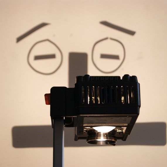 An overhead projector casts a shadow of a cartoonish, concerned face with raised eyebrows on a wall, creating a humorous and whimsical tone.