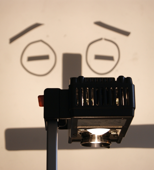 An overhead projector casts a shadow of a cartoonish, concerned face with raised eyebrows on a wall, creating a humorous and whimsical tone.