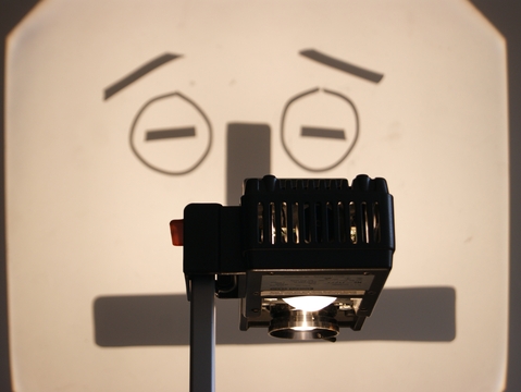 An overhead projector casts a shadow of a cartoonish, concerned face with raised eyebrows on a wall, creating a humorous and whimsical tone.