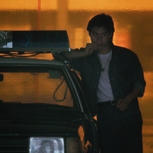 A still from As Tears Go By of a person leaning against a police car, set against a glowing orange background.