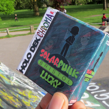A hand holds a Solarpunk-themed card game box labeled "Bristol" at a graffiti-covered skate park. People relax on grassy areas under sunny skies.