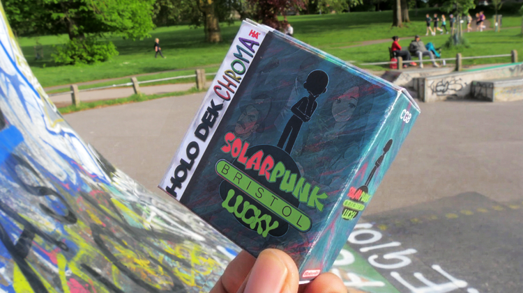 A hand holds a Solarpunk-themed card game box labeled "Bristol" at a graffiti-covered skate park. People relax on grassy areas under sunny skies.