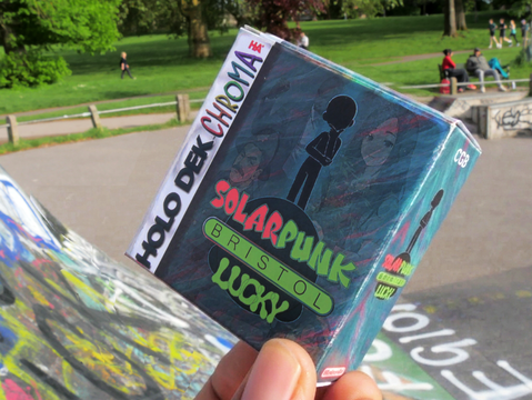 A hand holds a Solarpunk-themed card game box labeled "Bristol" at a graffiti-covered skate park. People relax on grassy areas under sunny skies.