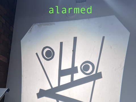 A projection on a wall shows abstract shapes resembling a surprised face. Above it, the word "alarmed" is written in green. The tone is humorous.