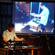 Ant Nasce a white man with glasses on stands at a Korg synthesizer, performing music under dim lighting. A large screen behind him displays a projected image of another person adjusting equipment, conveying a creative and focused atmosphere.