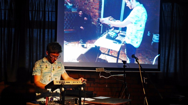 Ant Nasce a white man with glasses on stands at a Korg synthesizer, performing music under dim lighting. A large screen behind him displays a projected image of another person adjusting equipment, conveying a creative and focused atmosphere.