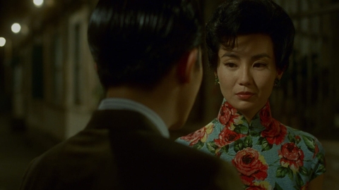 In still from In the Mood for Love of a man in a dark suit facing a woman wearing a blue floral qipao on a dimly lit street at night.