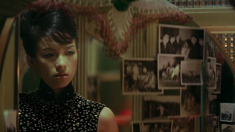 A still from 2046 of a person in a sleeveless, sparkly black top standing in front of a mirror decorated with various black-and-white photos.