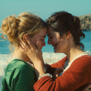 Two women in vintage dresses embracing on a sunny beach with a rocky cliff and ocean waves in the background.
